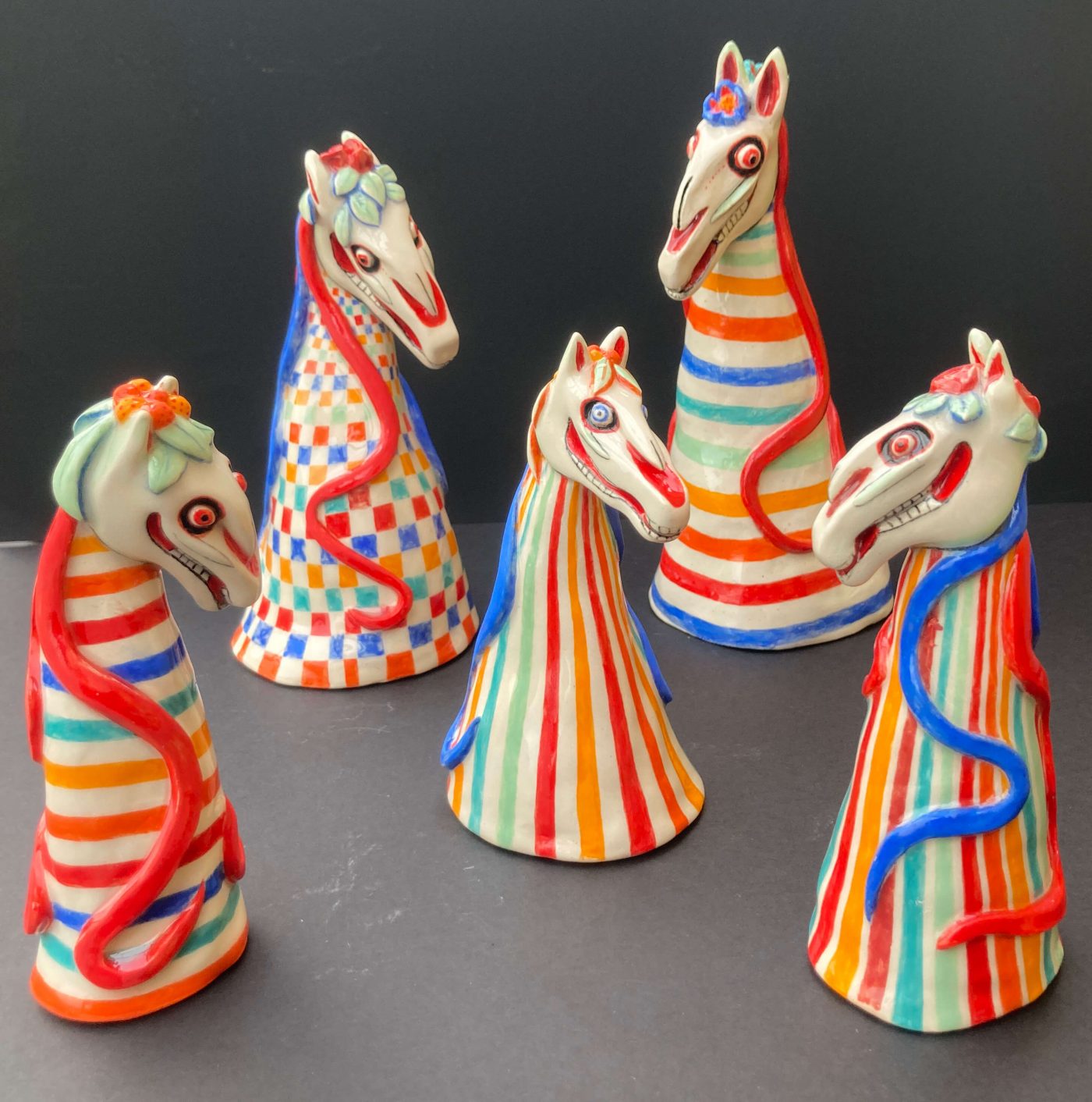 Five colorful ceramic figurines with horse skull heads and striped or checkered patterns stand on a dark surface, each adorned with decorative flowers and snakes.