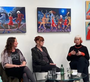 Three women sit and talk in front of colourful paintings during an indoor event; water glasses and a green bottle are on the table in front of them.